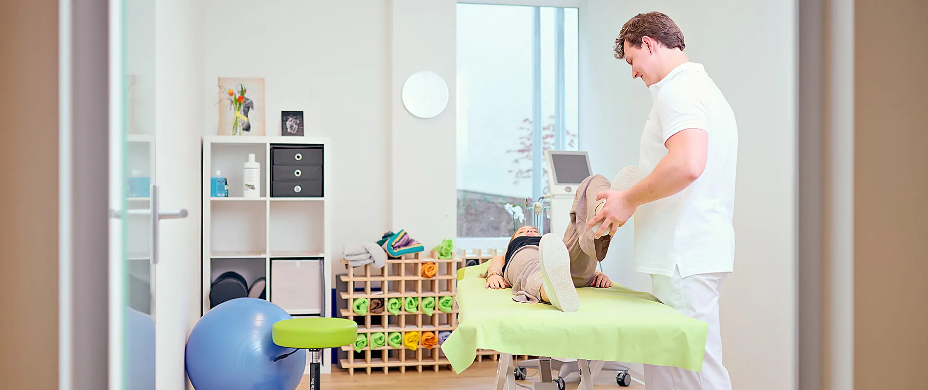 Ein Physiotherapeut hebt das gebeugte Bein einer Patientin an, die auf dem Rücken auf einer hellgrünen Behandlungsliege liegt. Im Hintergrund ein Regal mit gerollten Handtüchern und ein Fenster.