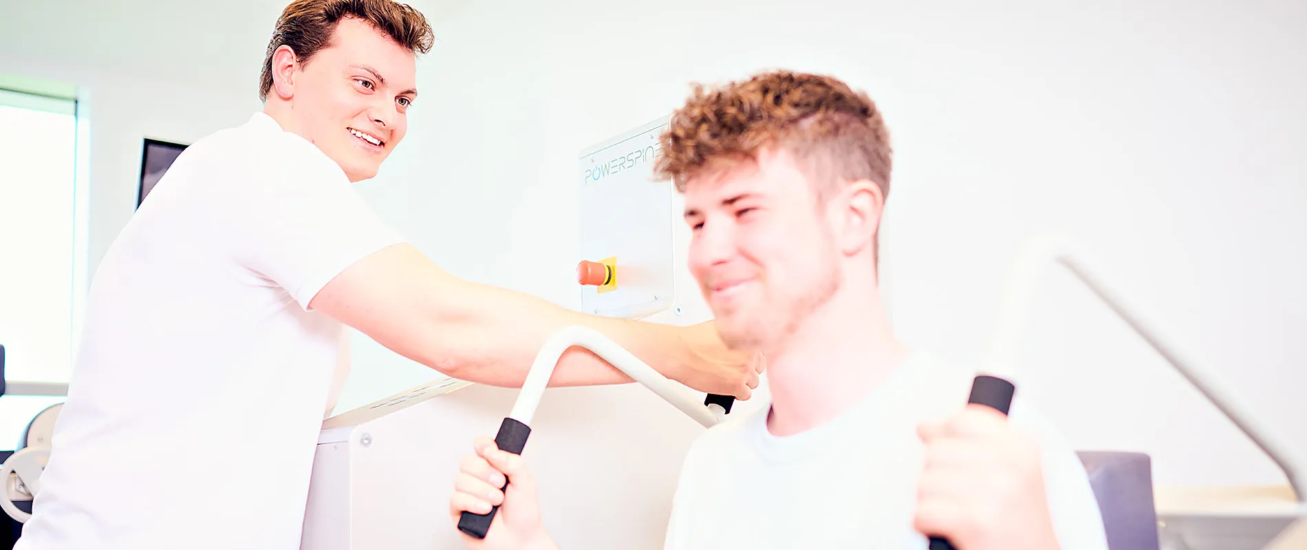 Ein Patient trainiert am POWERSPINE-Gerät, der Therapeut steht direkt neben ihm und korrigiert die Haltung
