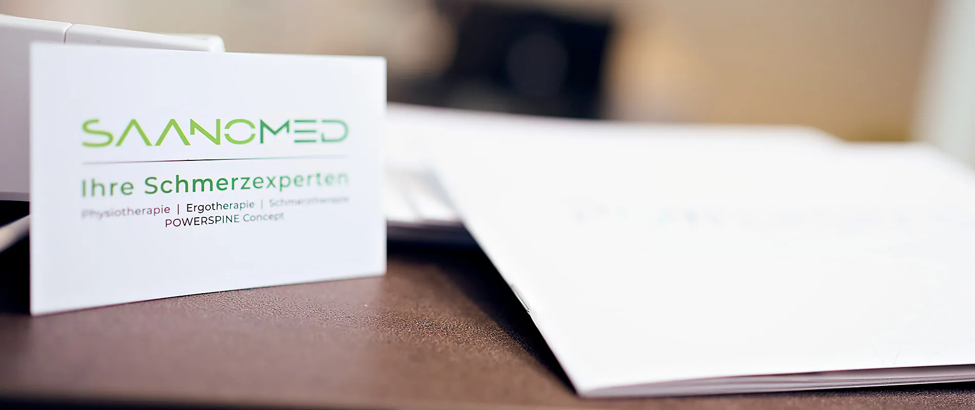 Eine Visitenkarte von SaanoMed mit Logo und dem Zusatz Ihre Schmerzexperten liegt auf dem Empfangstresen
